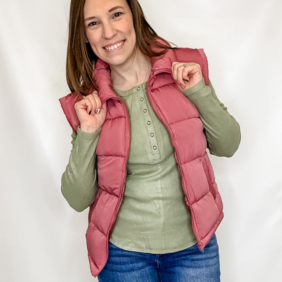 A person wearing a mauve pink puffer vest over a green shirt and blue jeans, posing against a white background.