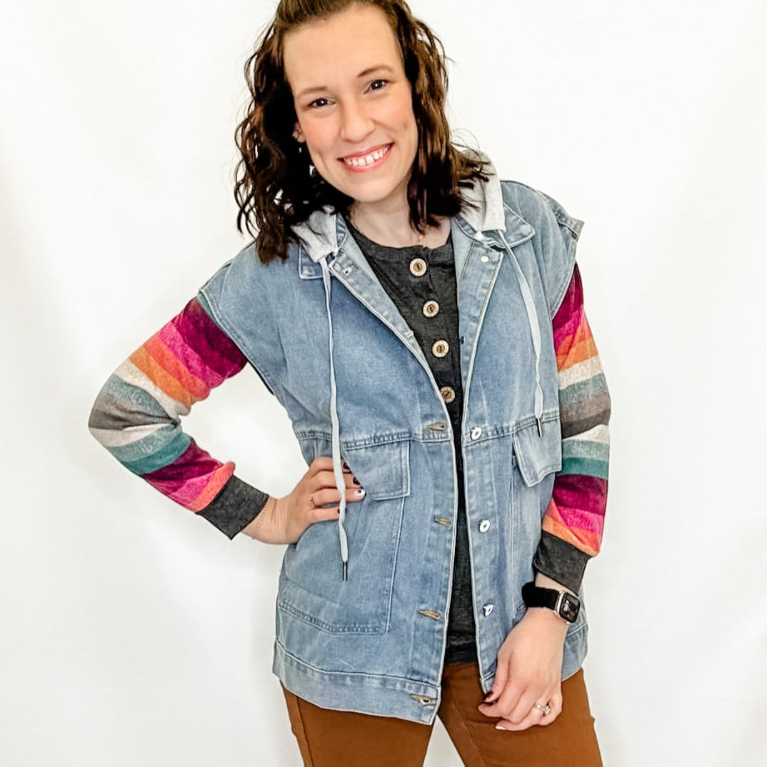 A woman wearing a hooded denim vest with multicolored stripes on the sleeves, paired with brown pants and standing against a white background.
